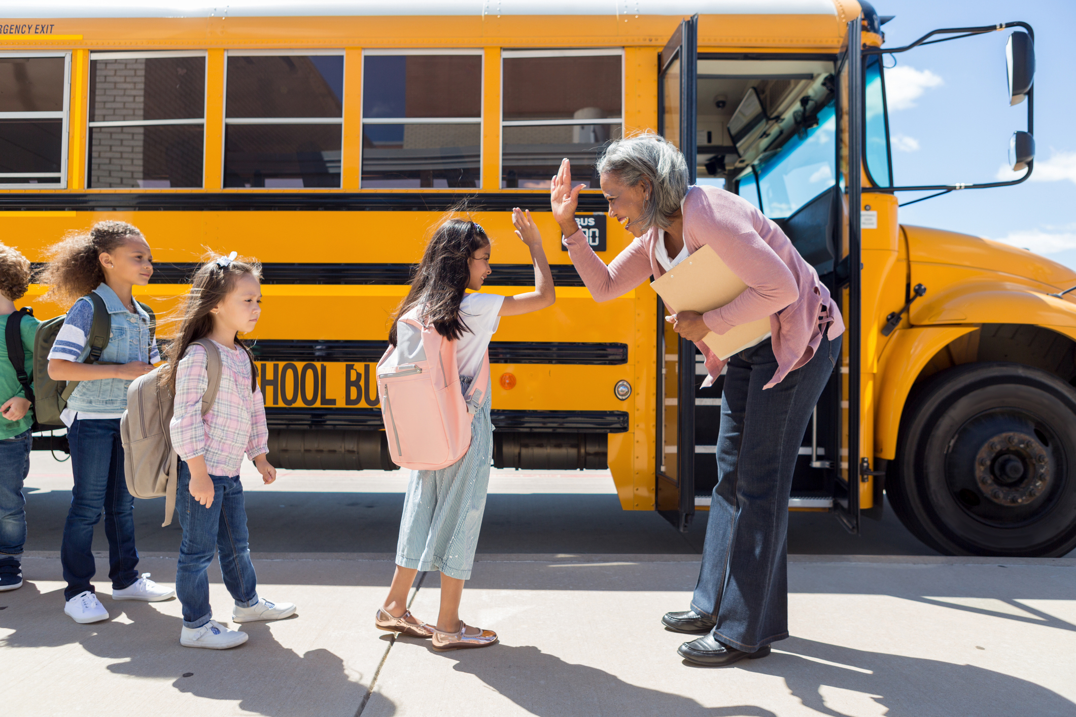 An education administrator stands outside of a school bus and gives student in line a high-five