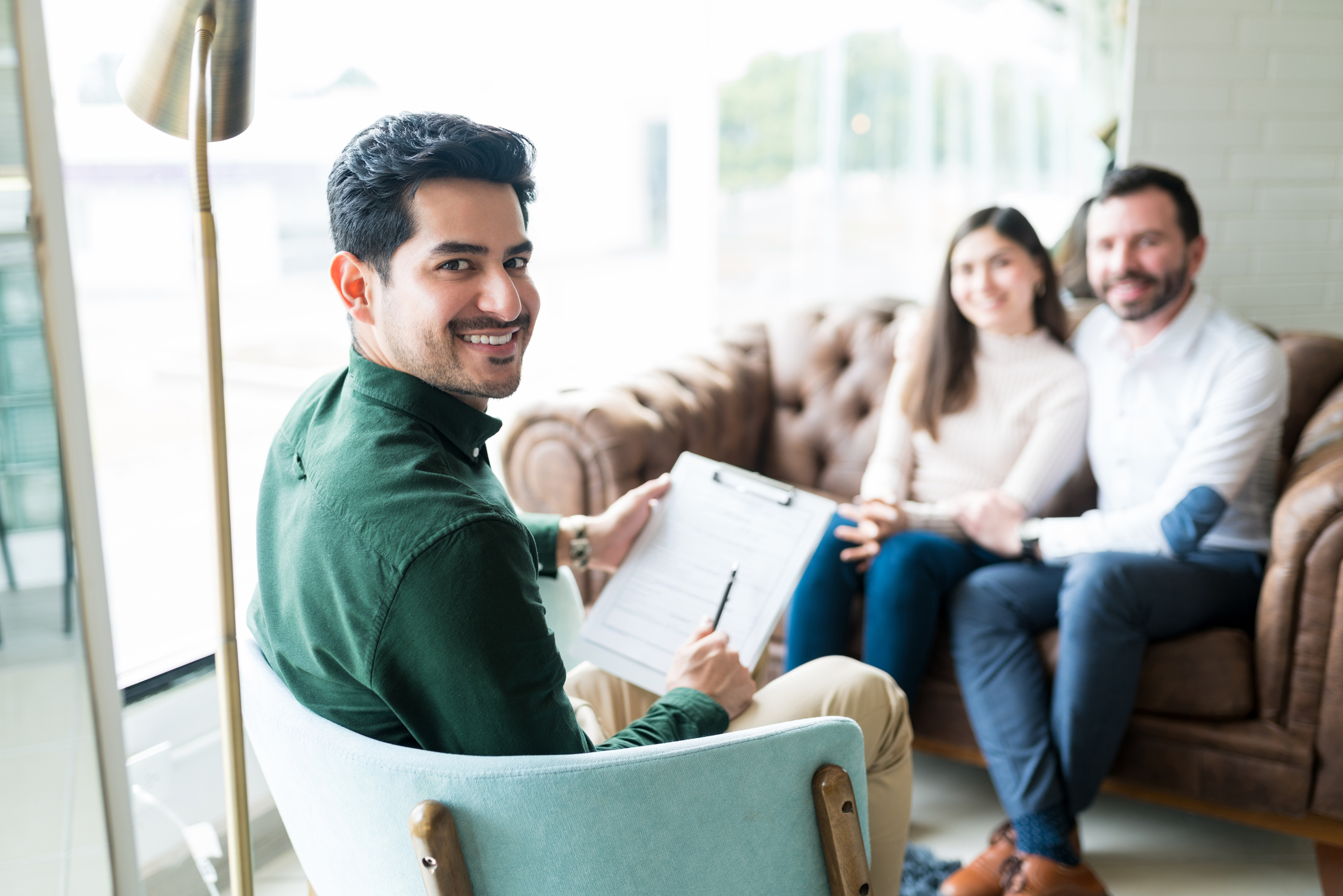 A Latino marriage and family therapist smiles at the camera while patients are seen in the background