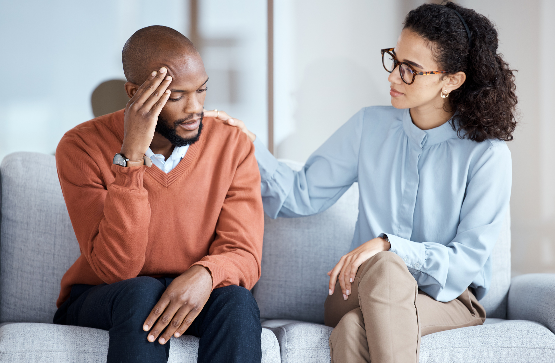 An African American man is seated on a couch while his African American female therapist consoles him