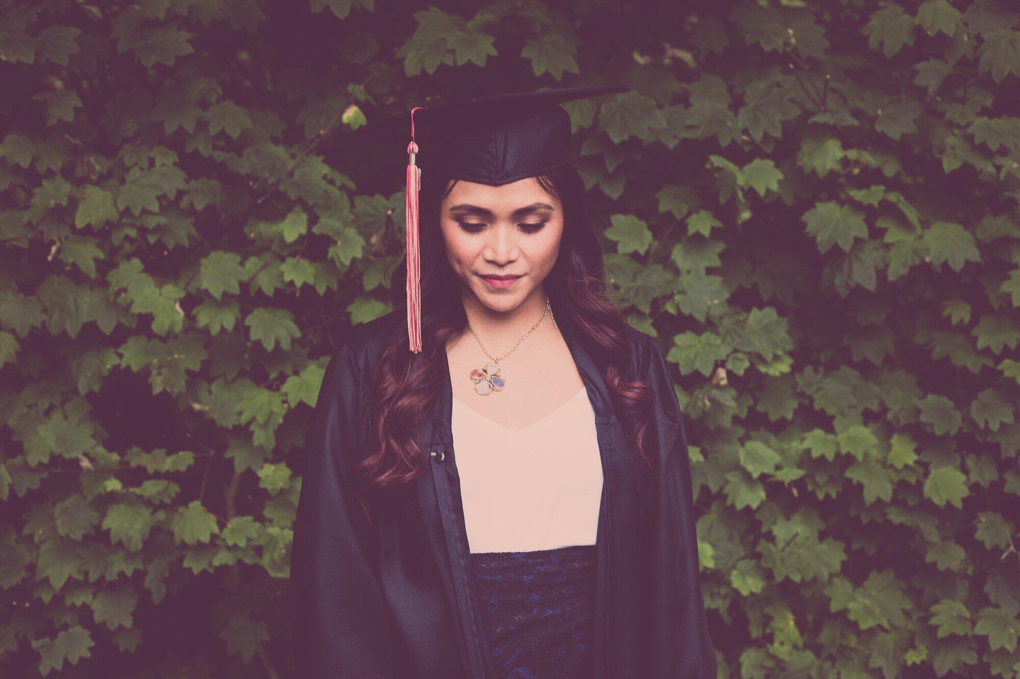 student in cap and gown standing in front of greenery