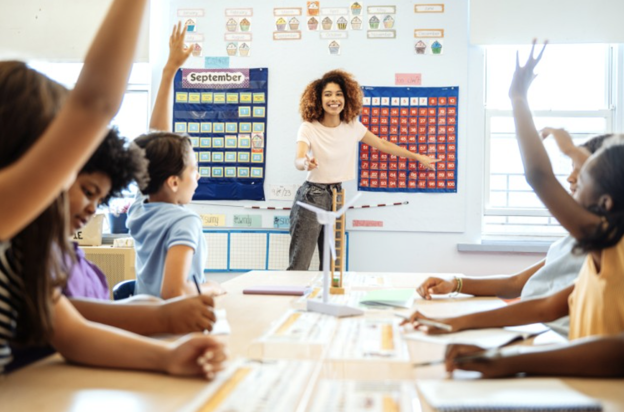 students raising their hands in a classroom with a teacher teaching 