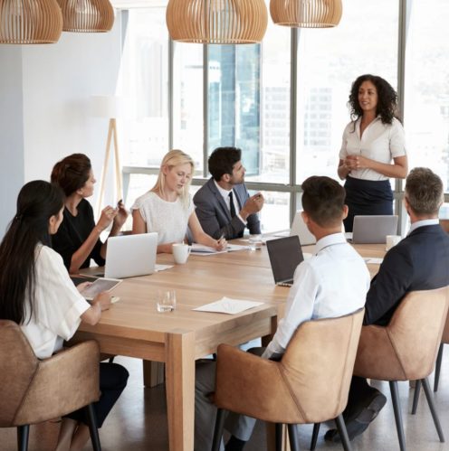 A business manager standing at the head of a desk leads a team meeting.